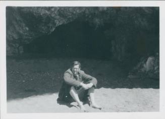 David Lewis sitting on the the beach with his elbows on his knees and his feet in the sand, with one of the caves fo the Grotte Marine di Capo Palinuro behind him.