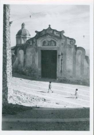 A church with big square doors and an angelic marble relief set in the facade above it. A small domed tower rises to the left behind it and two very small children play out in front.
