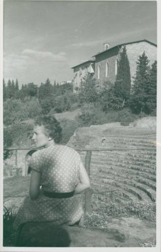 WBG sitting at the top of a Roman ampitheatre in Fiesole, wearing a polkadot dress and looking away from the camera. A building stands in the background