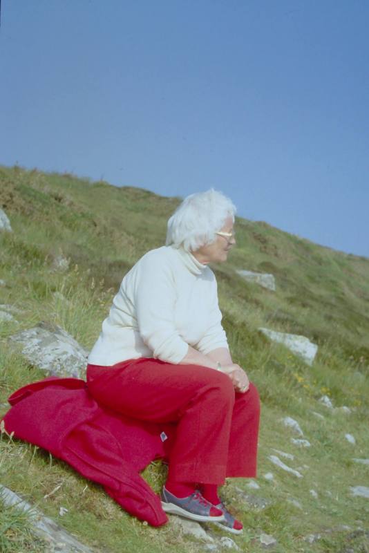 6 images of Wilhelmina Barns-Graham sitting on grassy hill. Red jacket and trousers.