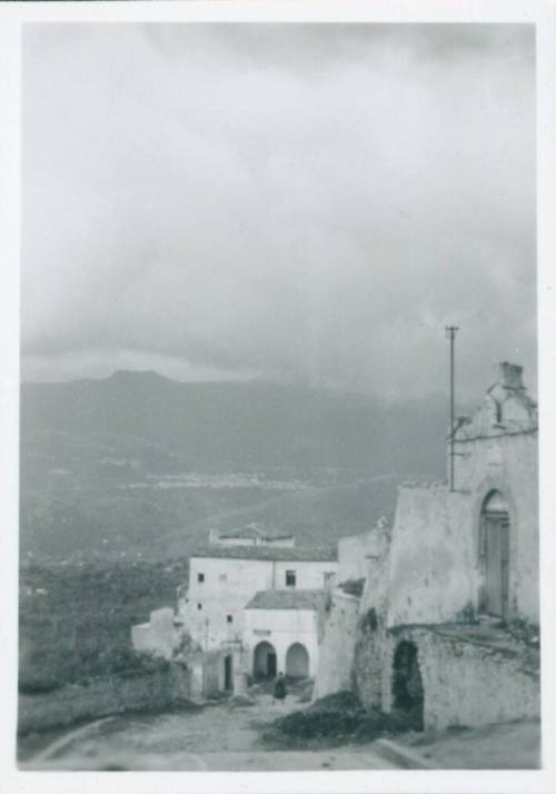 Sicilian street with buildings to the right and in front of the road. The mountains stretch out in the background and it is a cloudy day.