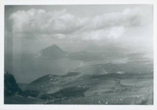 Countryside with farmland to the right and the ocean to the left. A hill forms a small peninsula in the left background