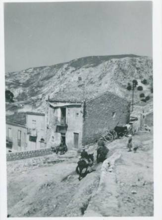 Villagers going about their business. Three men are riding donkeys in the center of the frame while a woman watches with her dog. Houses and a hill with sparse vegetation rise in the background.