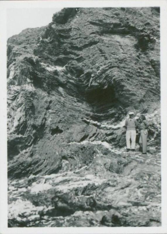 A woman and man stand next to one another against a rock face. They are both small in the fram, engulfed by the rock.