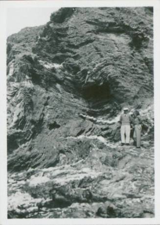 A woman and man stand next to one another against a rock face. They are both small in the fram, engulfed by the rock.