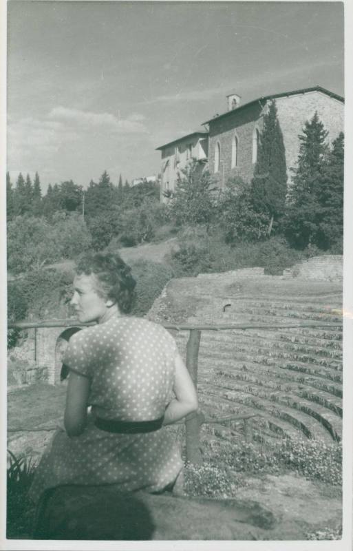 WBG sitting at the top of a Roman ampitheatre in Fiesole, wearing a polkadot dress and looking away from the camera. A building stands in the background
